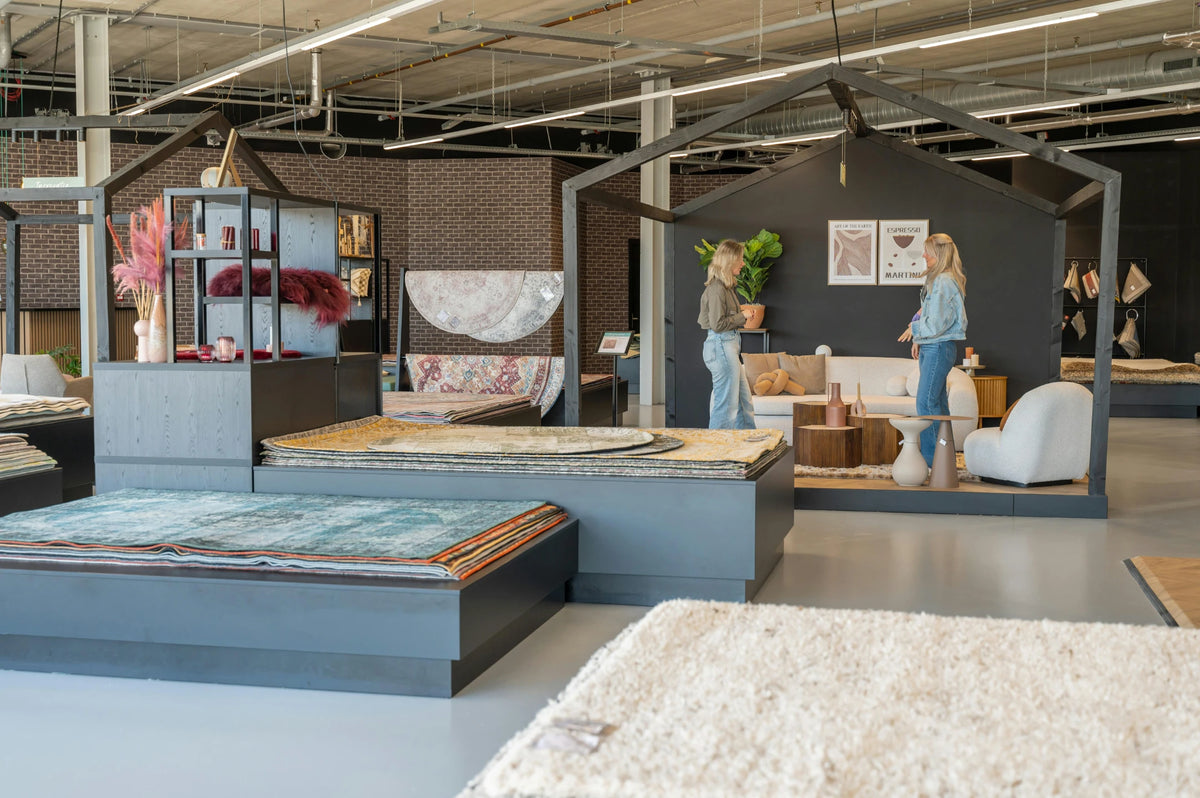 Two visitors talking in a styled living setting inside the Volero Amsterdam showroom, surrounded by stacked rugs on display platforms